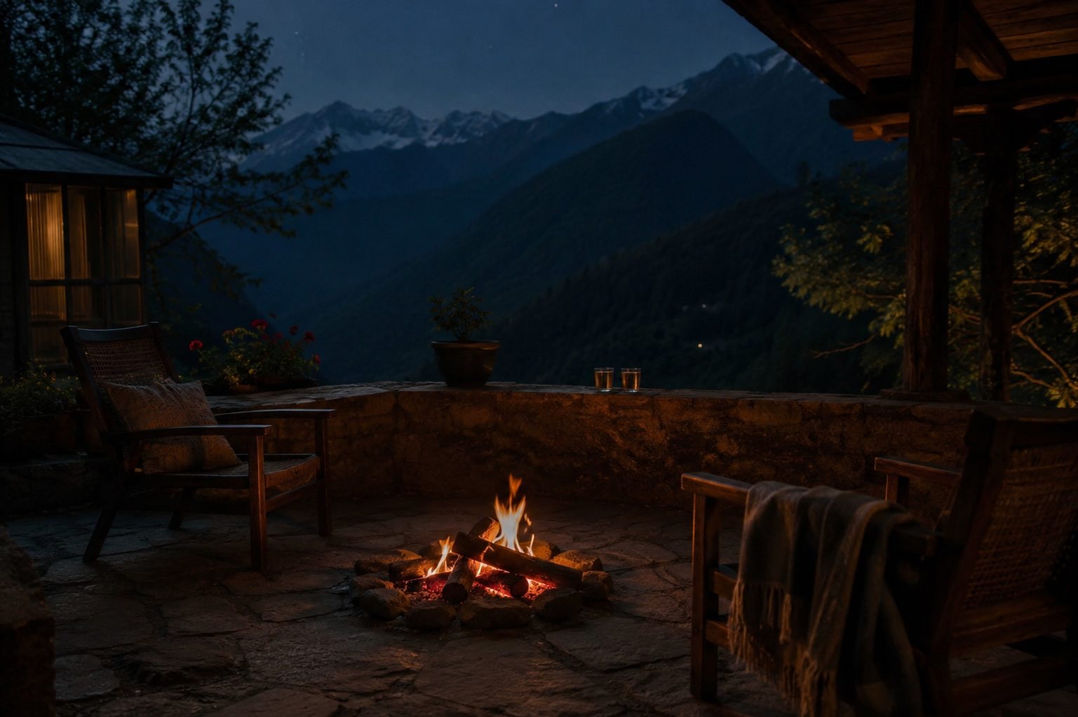 A small evening fire in a stone courtyard, two chairs and a blanket beside it, tea glasses on the parapet, the dark valley and snowline beyond.