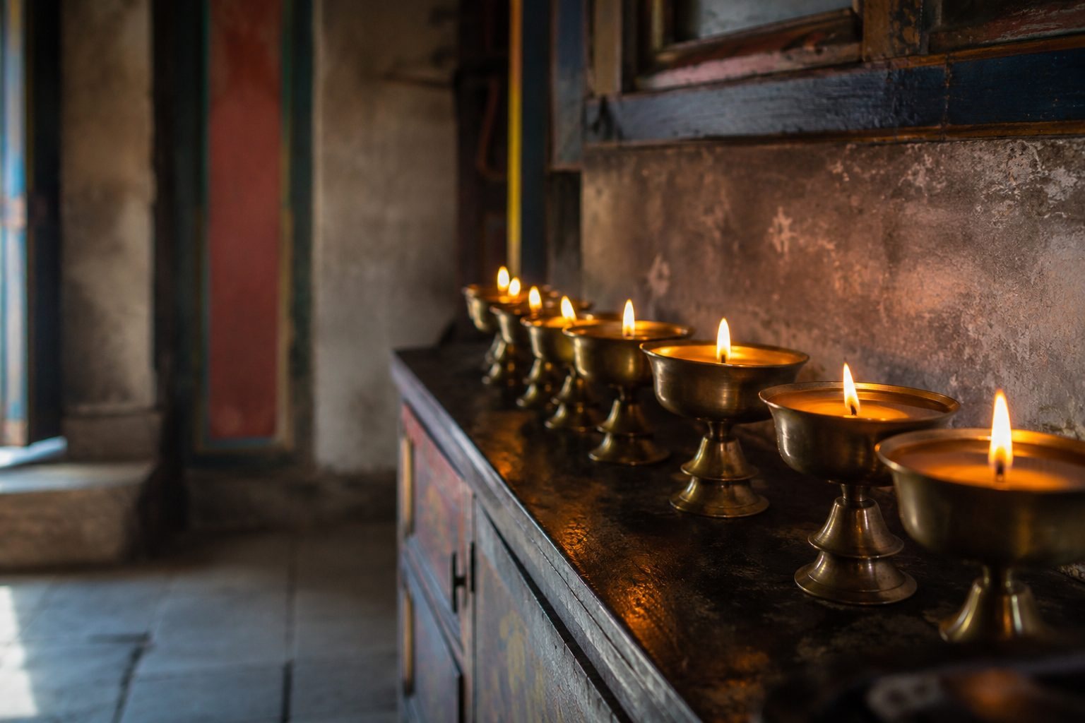 A row of brass butter lamps burning on a dark wooden chest in a quiet, weathered interior, morning light from a side window.