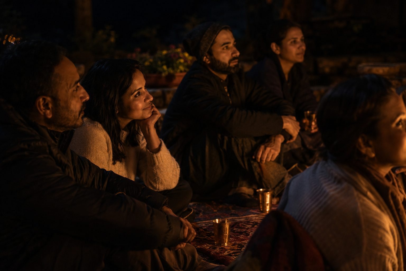 Guests seated on rugs by firelight, warm glasses in hand, listening — faces turned toward the song.