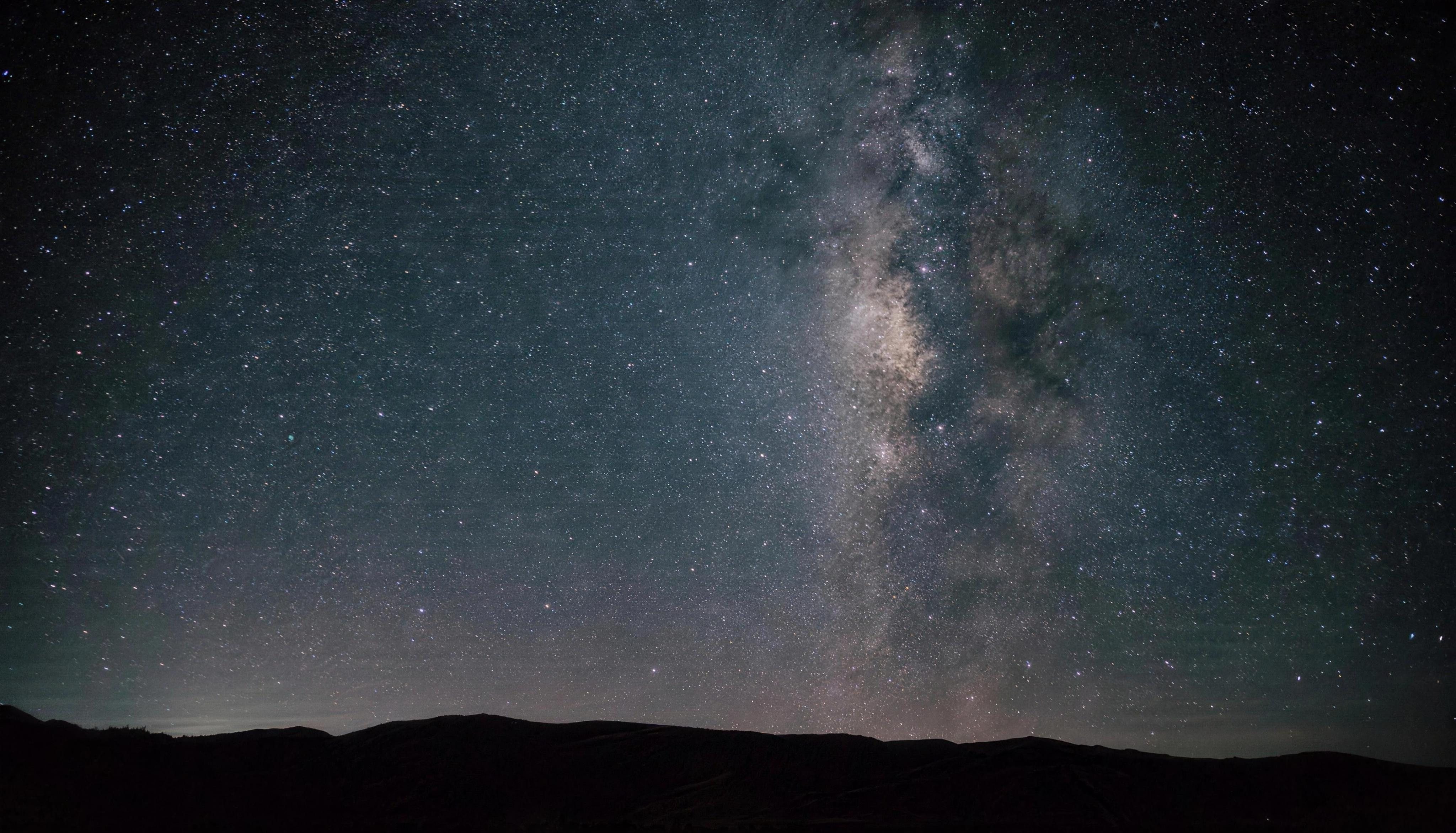 A deep night sky over a dark ridge, the Milky Way standing above the mountains and a faint warm glow along the horizon.