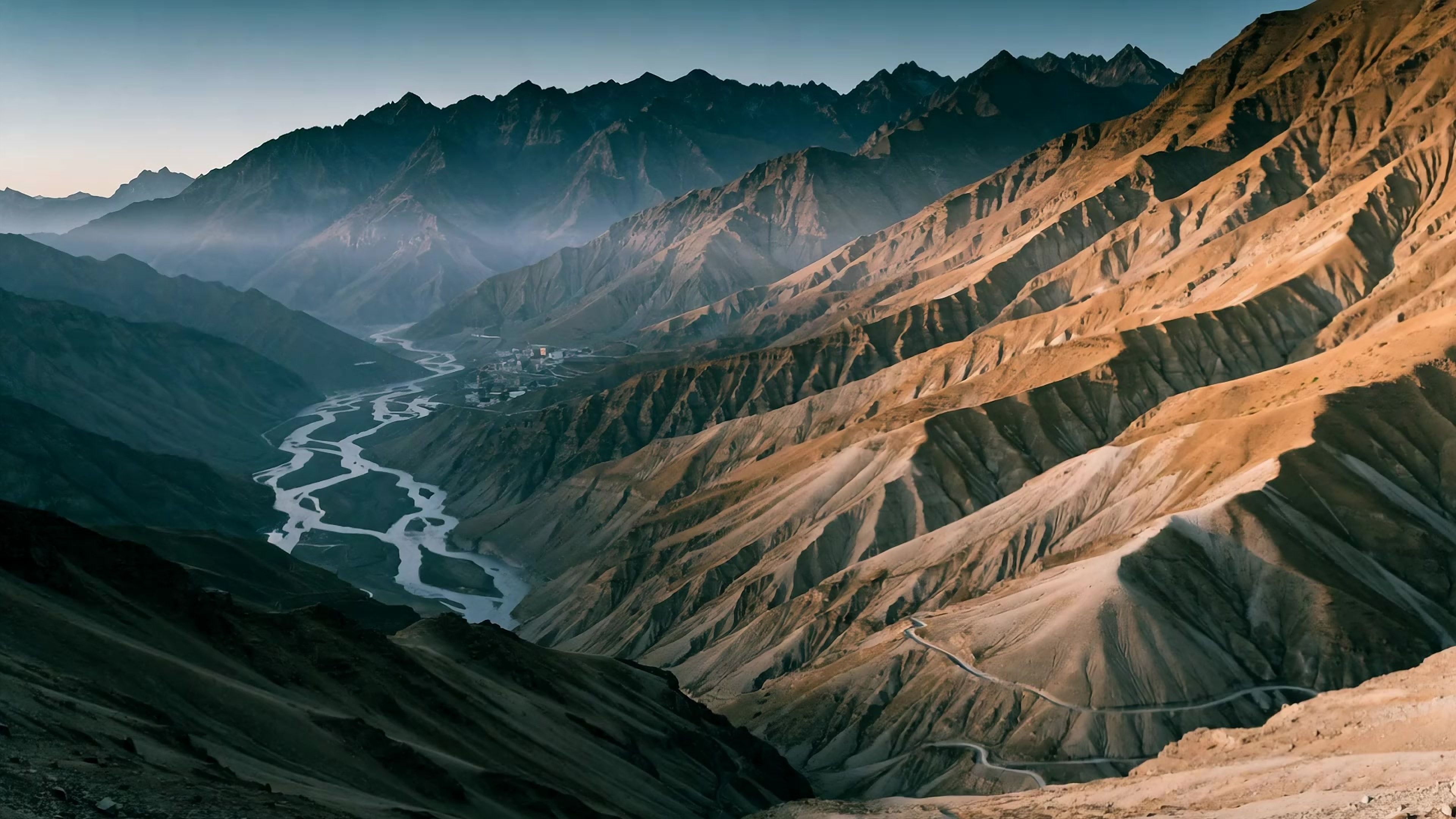 A wide Spiti river valley at dusk: a braided river threading the valley floor, layered desert ridges fading into blue haze, a small village and a thread of road on the far slopes.